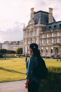 side view photo of standing woman in black cap and blue denim shirt carrying backpack with building in the background